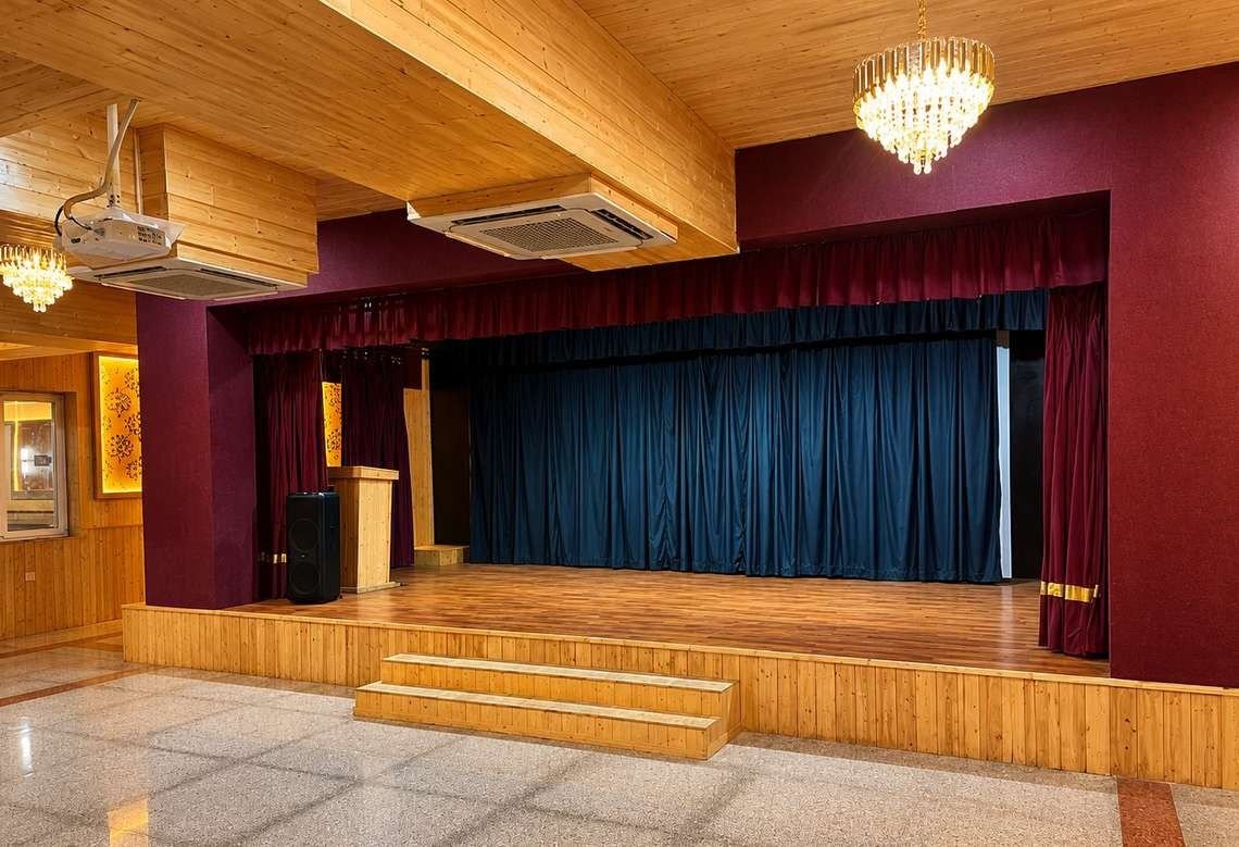 Stage hall at AIS Residency with a raised wooden stage and steps, maroon proscenium walls, blue stage curtains and a chandelier